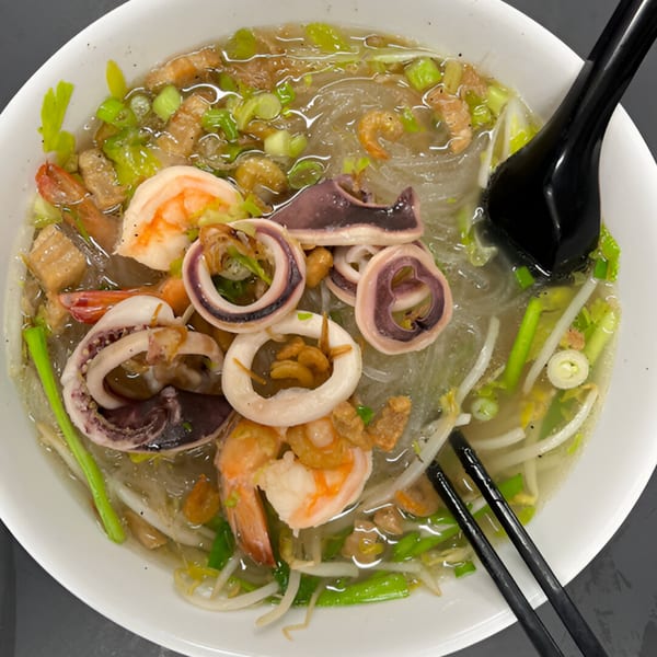 Seafood noodle soup with shrimp, squid, green onions, and bean sprouts in a white bowl.