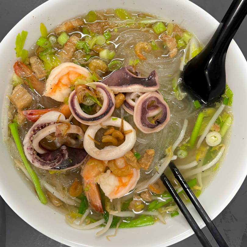 Seafood noodle soup with shrimp, squid, green onions, and bean sprouts in a white bowl.